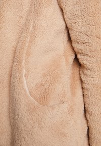 Tissu en polaire beige pelucheux avec une couture visible et un détail de poche, montrant une surface douce et texturée.