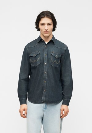 Jeune homme aux cheveux mi-longs et foncés portant une chemise en jean foncé à boutons avec poches avant et un jean bleu clair, debout dans une posture neutre.
