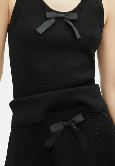 Haut en tricot sans manches noir et jupe avec des nœuds en satin noirs assortis à la poitrine et à la taille, portés par une personne.