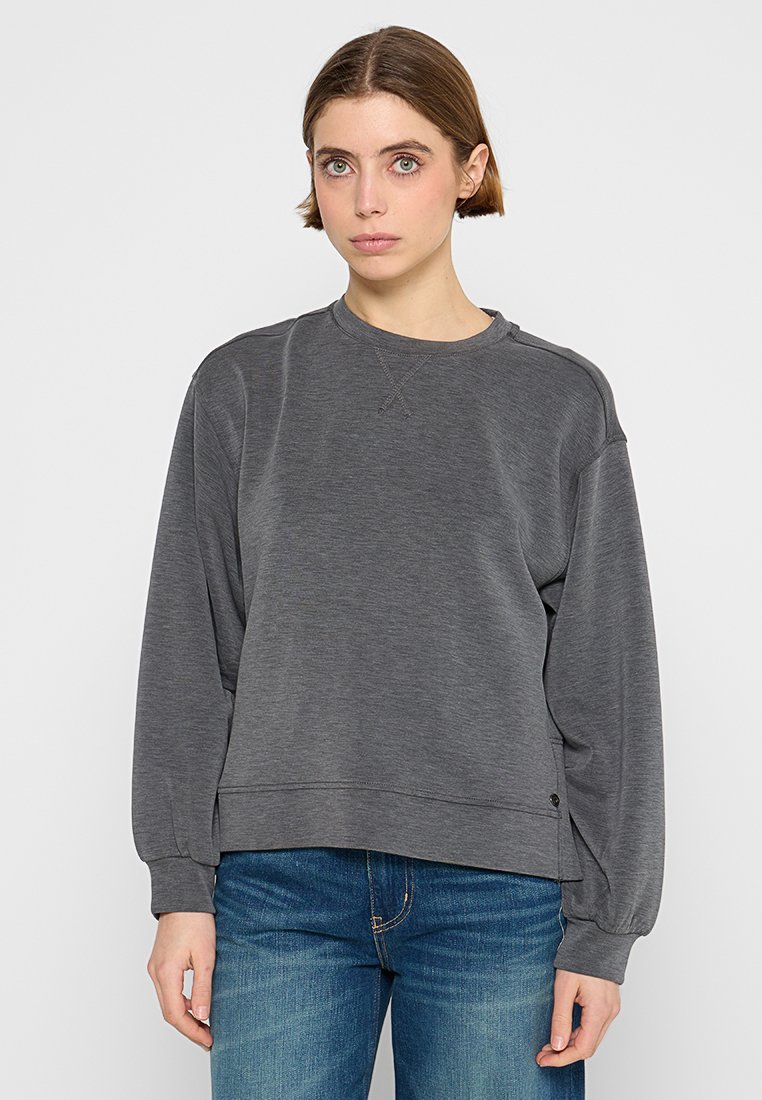 Frau trägt einen dunkelgrauen Langarm-Sweatshirt und blaue Jeans, steht vor einem schlichten hellen Hintergrund.