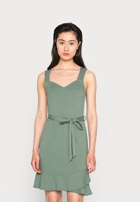 Vestido verde menta com corpete justo, alças largas e decote em V. Apresenta uma cintura atada e uma bainha em babados evasê na saia. Textura suave.