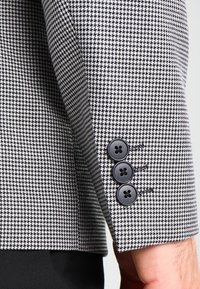 Zblizenie na rękaw czarno-białego żakietu w kratę houndstooth z trzema czarnymi guzikami na nadgarstku osoby oraz czarnymi spodniami.