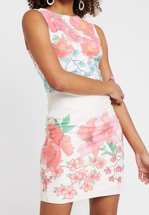 Robe blanche sans manches avec de grands motifs floraux roses et corail portée par une personne aux cheveux bouclés et portant des boucles d'oreilles blanches circulaires.