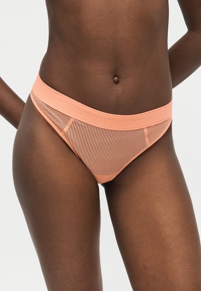 Γυναίκα που φοράει μαγιό στιλ εσώρουχο ροδάκινου με ρίγες και το λογότυπο "Polo Ralph Lauren" στη μέση, στέκεται μπροστά σε απλό φόντο.