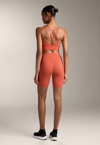 Sportos szett meleg korall színben: crisscross pántos sportmelltartó, magas derekú rövidnadrág és fekete sportcipő. Sima textúra, testhezálló fazon.