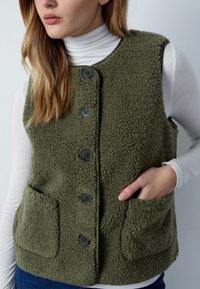 Frau, die eine olivgrüne Fleece-Weste mit Knöpfen und Fronttaschen über einem weißen Rollkragenpullover trägt, mit einer Hand in einer Tasche.