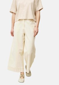 Top court beige avec une finition texturée, associé à un pantalon large crème clair avec un ourlet brut. Des chaussures dans des tons neutres complètent la tenue.