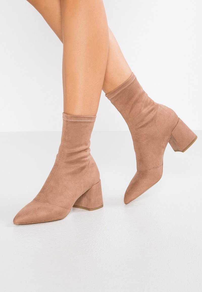 Bottines en suède beige clair avec bout pointu, tige ajustée, finition texturée et talon carré robuste. Détail de coutures visibles.