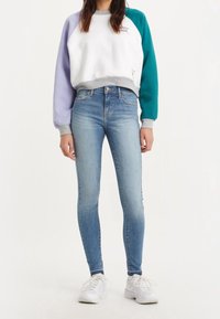 Kvinna klädd i en vit sweatshirt med lila och teal ärmar, ljusblå tajta jeans och vita sneakers, stående mot en vit bakgrund.