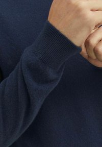 Pulover din lână bleu marin, cu manșete încrețite, croială ajustată, textură moale și o ușoară strălucire, evidențiind calitatea materialului și detalii de construcție.