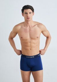 Bóxer azul marino con un logo de Lacoste en la cinturilla negra, que presenta un ajuste ceñido y una textura de tela suave. Sin patrones visibles.