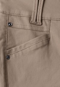Gros plan d'une poche de pantalon en tissu beige avec des coutures visibles et deux rivets métalliques près de l'ouverture de la poche et des passants de ceinture au-dessus.