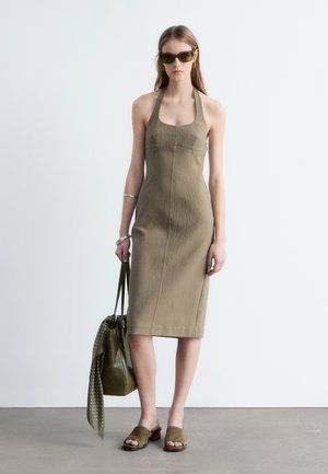 Kvinde iført en ærmeløs olivengrøn midi-kjole, solbriller, slidesandaler, sølvarmbånd, holder en matchende taske med et prikket tørklæde.