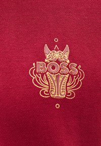 Κόκκινο ύφασμα με υφή, που διαθέτει ανάγλυφο χρυσό λογότυπο της μάρκας "BOSS" με περίτεχνα διακοσμητικά στοιχεία γύρω του.