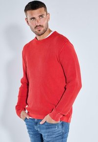 Red ribbed long-sleeve sweater with a round neckline and relaxed fit. Features textured fabric and ribbed cuffs. Paired with blue denim jeans.