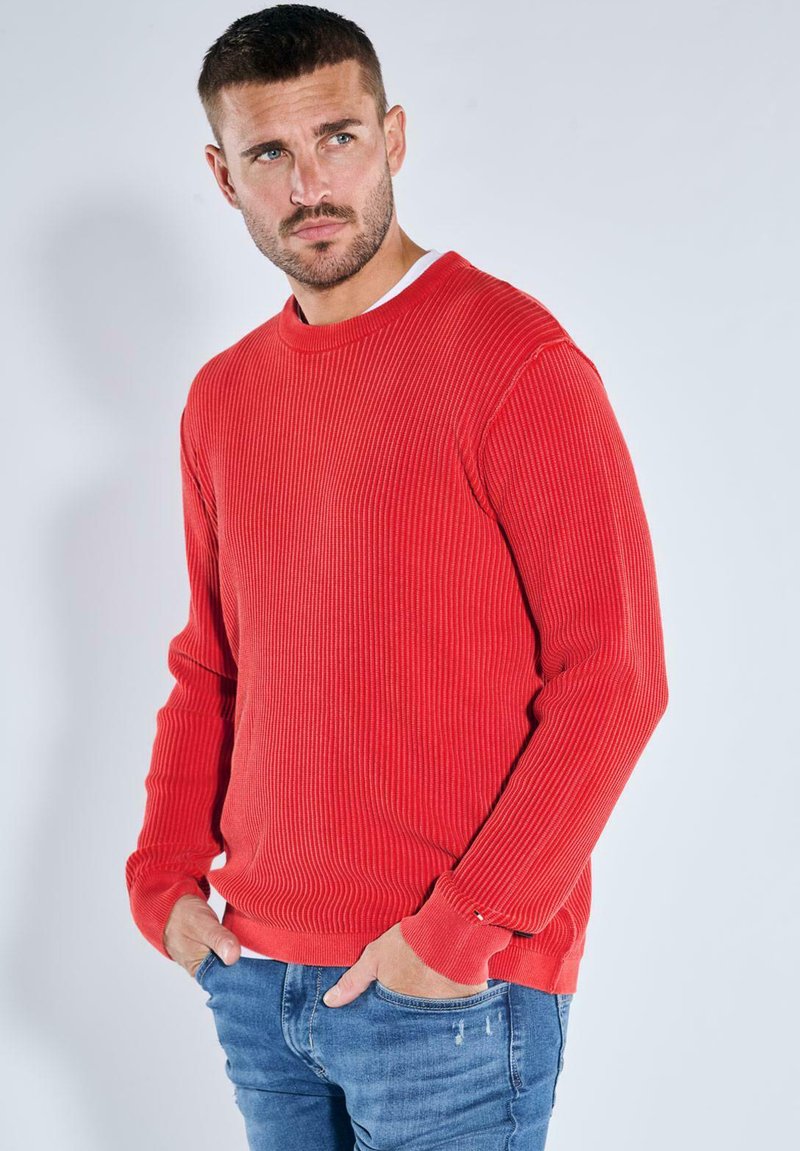 Red ribbed long-sleeve sweater with a round neckline and relaxed fit. Features textured fabric and ribbed cuffs. Paired with blue denim jeans.