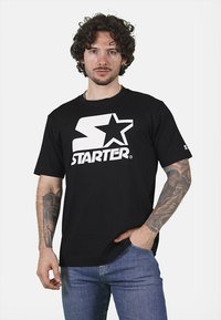 T-shirt in cotone nero con un grande logo bianco a forma di "S" e stella, maniche corte e vestibilità ampia. Indossato con jeans in denim blu.