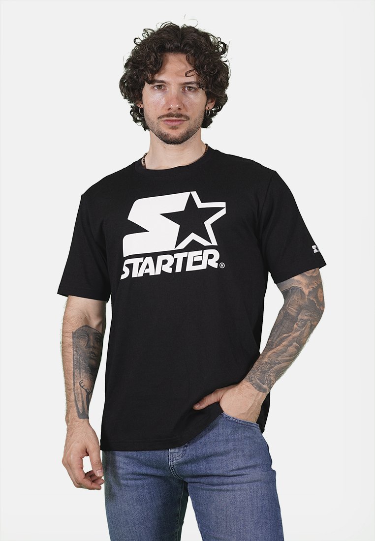 T-shirt in cotone nero con un grande logo bianco a forma di "S" e stella, maniche corte e vestibilità ampia. Indossato con jeans in denim blu.