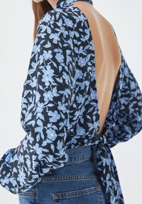 Blouse à imprimé floral bleu marine avec fleurs bleu clair, manches longues, découpe profonde dans le dos et détail de taille nouée. Tissu texturé et léger.