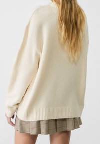 Kvinna med långt blont hår som bär en lös krämfärgad tröja och en veckad beige rutig kjol, sedd bakifrån mot en enfärgad bakgrund.