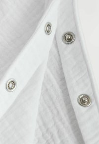 Gros plan d’un tissu blanc avec des boutons-pression métalliques partiellement ouverts, montrant des couches de tissu texturé en dessous.