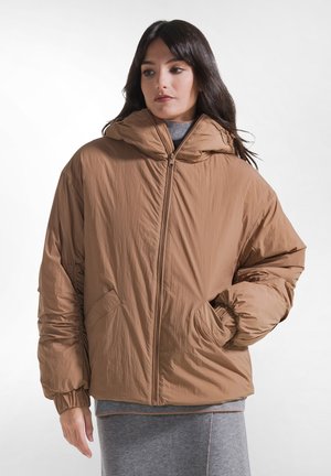 Jeune femme portant une doudoune beige à capuche avec une fermeture éclair devant et les mains dans les poches, debout devant un fond blanc uni.