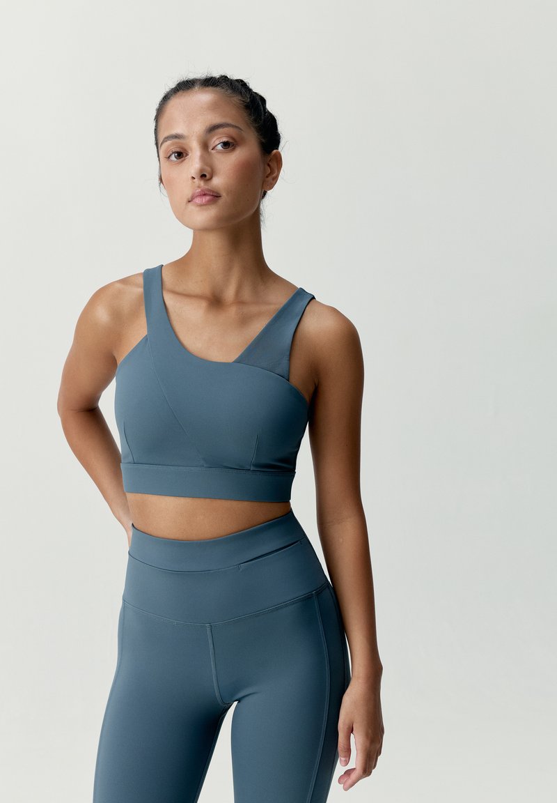 Modrý športový crop top s asymetrickým dizajnom a širokými ramienkami, doplnený o vysoké legíny v zodpovedajúcej farbe a hladkej textúre.