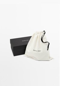 Sac en coton avec cordon de serrage en crème et logo noir, placé à côté d'une boîte rectangulaire noire mat. Les deux articles sont soigneusement disposés sur un fond blanc.