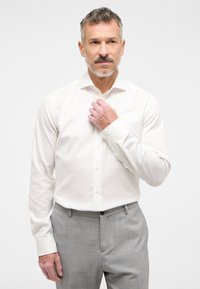 Weißes Langarmhemd mit klassischem Kragen, Knopfverschluss und Manschetten, kombiniert mit grauen Hosen, auf einem neutralen Hintergrund.