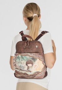 Brauner Rucksack mit einer fantasievollen Illustration eines Kindes und dem Satz "Der Schlag des Herzens der Erde", mit dekorativen Reißverschlussanhängern.