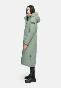 Grüner, gepolsterter langer Mantel mit Kapuze, vorderen Knöpfen, seitlichen Taschen und geprägtem Schulterpatch. Kombiniert mit schwarzen Stiefeln und dunkler Hose.