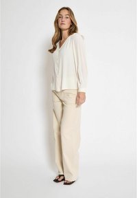 Jasno beżowa bluzka z długimi rękawami, zapięciem na guziki i dekoltem w kształcie litery V. Połączona z kremowymi spodniami o szerokich nogawkach i czarnymi sandałami. Gładka tekstura materiału.