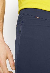 Gros plan sur une personne portant un pantalon bleu marine avec une poche zippée verticale et une chemise jaune rentrée.