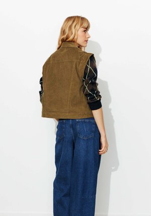 Gilet en velours côtelé marron à col montant, associé à un pull noir et vert à motifs et un pantalon en denim bleu ample.