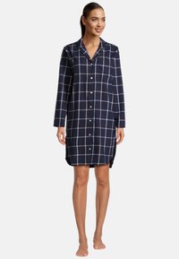 Donna in piedi a piedi nudi che indossa una camicia da notte blu navy con bottoni, decorata con quadretti bianchi e grigi.