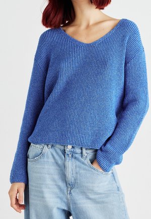 Personne aux cheveux roux portant un pull tricoté bleu à manches longues et un jean en denim bleu clair, main dans la poche, sur un fond uni.