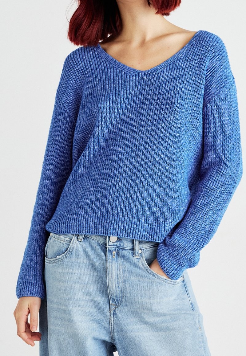Personne aux cheveux roux portant un pull tricoté bleu à manches longues et un jean en denim bleu clair, main dans la poche, sur un fond uni.