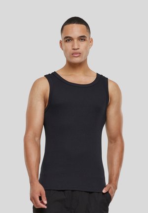Junger Mann mit kurzen Haaren, der ein schwarzes ärmelloses geripptes Tanktop und schwarze Hose trägt, vor einem einfarbigen hellgrauen Hintergrund stehend.