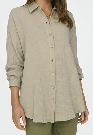Chemise ample beige texturée à boutons, manches longues retroussées aux poignets, portée avec un pantalon vert olive, visible de la poitrine jusqu'aux hanches.