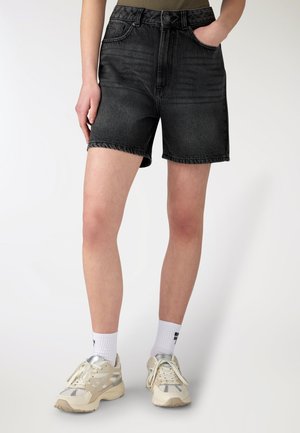 Persona che indossa pantaloncini di jeans neri, calzini bianchi con scritta e scarpe da corsa beige, in piedi contro uno sfondo chiaro e uniforme.