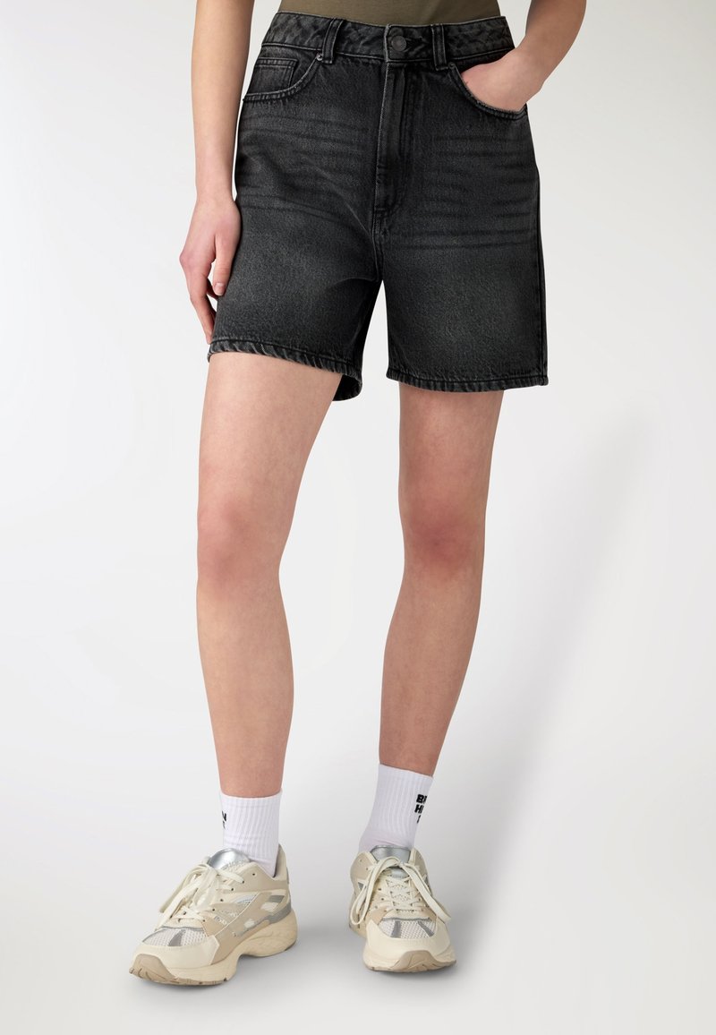 Persona che indossa pantaloncini di jeans neri, calzini bianchi con scritta e scarpe da corsa beige, in piedi contro uno sfondo chiaro e uniforme.