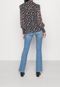 Floral-patterned, sheer black blouse with ruffled shoulders and button closure at the back, paired with light blue flared jeans and black boots.