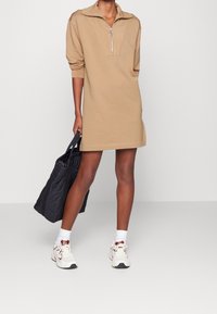 Beige, langärmliges Zip-Pulloverkleid mit hohem Kragen und Ring-Zipper. Getragen mit weißen Turnschuhen und einer schwarzen Tragetasche.
