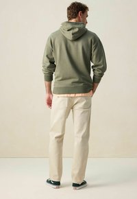 Man staat met zijn rug naar ons gekeerd, draagt een olijfgroene hoodie, lichtbeige broek, perzikkleurig shirt eronder en zwarte lage sneakers met witte zolen.