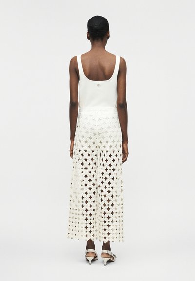 Femme vue de dos portant un haut blanc sans manches et un pantalon large en dentelle florale blanche, avec des sandales à talons, sur un fond uni.