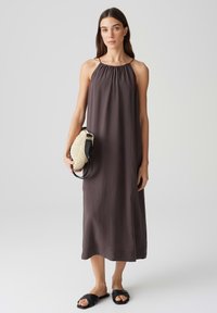 Dunkelbraunes, ärmelloses Maxikleid mit gerafftem Ausschnitt, glatter Textur, kombiniert mit einer hellen, gewebten Handtasche und schwarzen Sandalen.