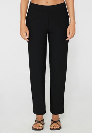 Donna che indossa pantaloni neri a gamba dritta e sandali neri con anelli d’oro sulle dita dei piedi, in piedi davanti a uno sfondo grigio chiaro uniforme.