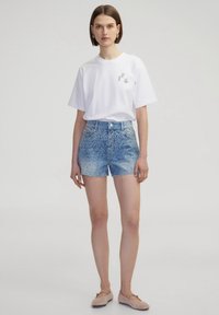 Camiseta blanca de manga corta con un peque�ño logo gris, combinada con pantalones cortos de mezclilla azul claro que presentan un acabado en degradado y detalles de tachuelas.