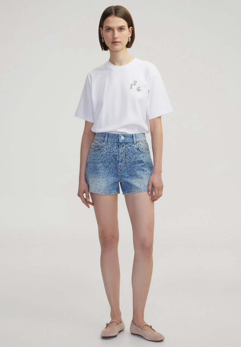 Witte t-shirt met korte mouwen en een klein grijs logo, gecombineerd met lichtblauwe denim shorts met een gradatieafwerking en studs.