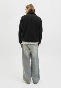 Person med skulderlangt hår iført en sort jakke, ternet skjorte, løse lys blå jeans og hvide sko, der vender bort fra kameraet på en ensfarvet baggrund.